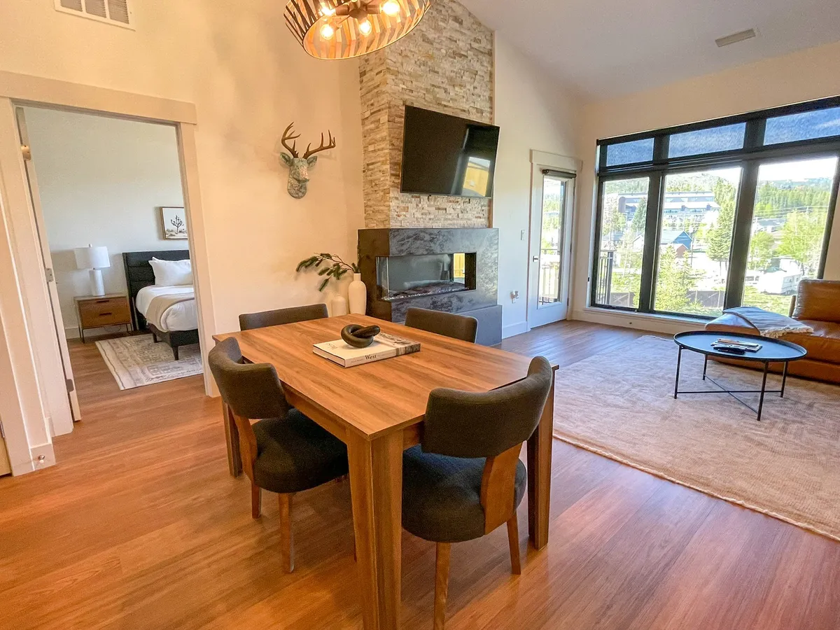 A cozy and contemporary interior featuring a dining table set near large windows, with a fireplace, leather sofa, and decorative accents. The space is filled with natural light, and the arrangement is ideal for dining, relaxing, or unwinding by the fire after a day outdoors.