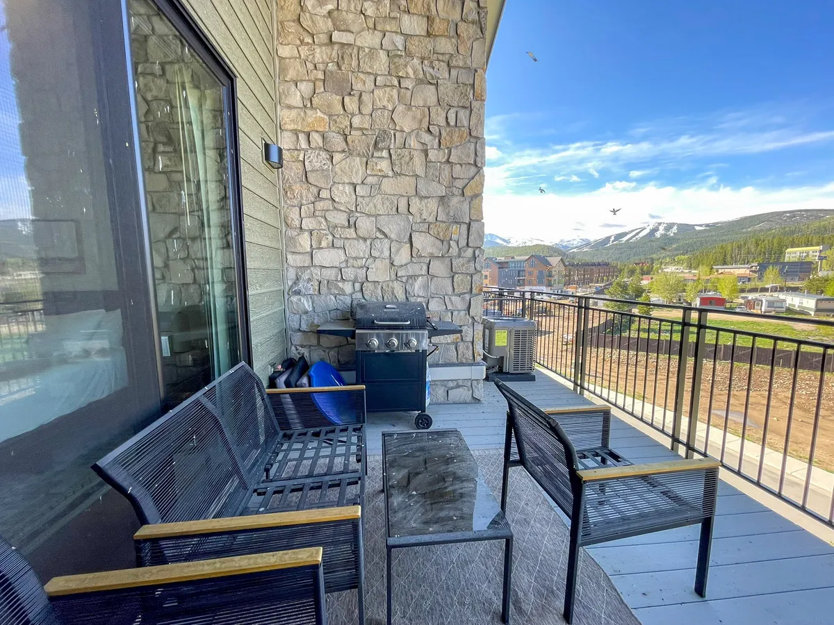 This image features a stone-walled balcony or patio area with a natural gas grill, comfortable seating, and an expansive mountain view in the background. The relaxed atmosphere invites guests to imagine barbecuing or unwinding outdoors.