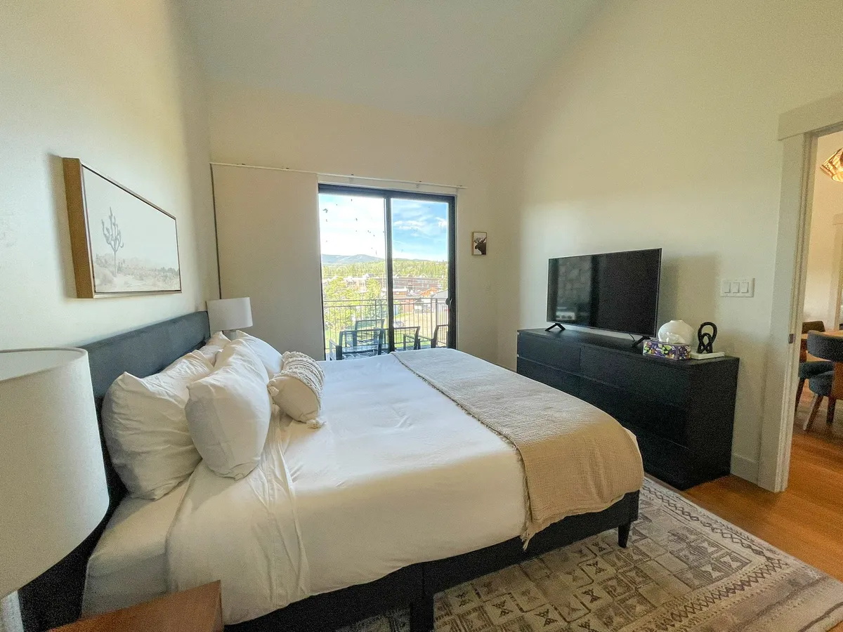 A cozy bedroom featuring a comfortable bed, wall-mounted television, window with scenic view, nightstand, and decorative artwork. The setting suggests a restful, private environment perfect for winding down.