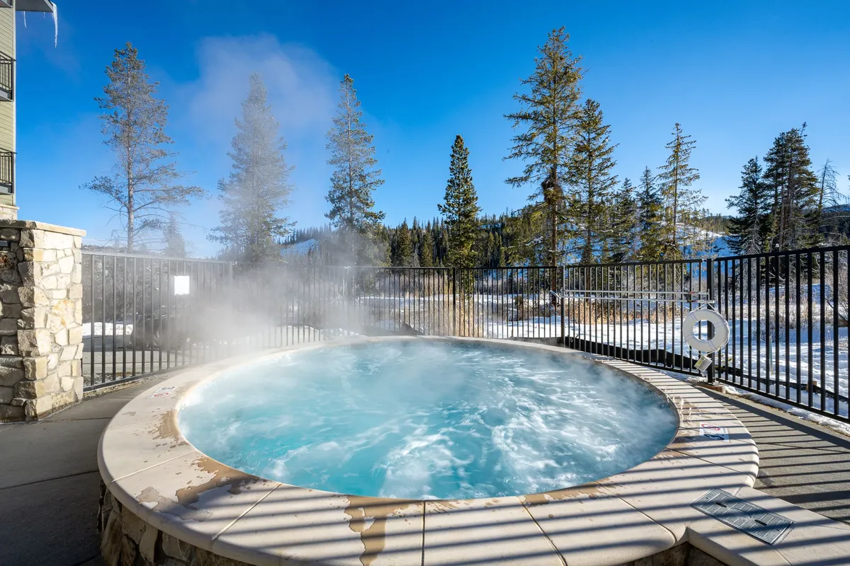 A serene outdoor hot tub is tucked among snowy trees, with the mountains visible in the background. The soothing setting is perfect for relaxing after a day of adventure, especially in the cooler months.