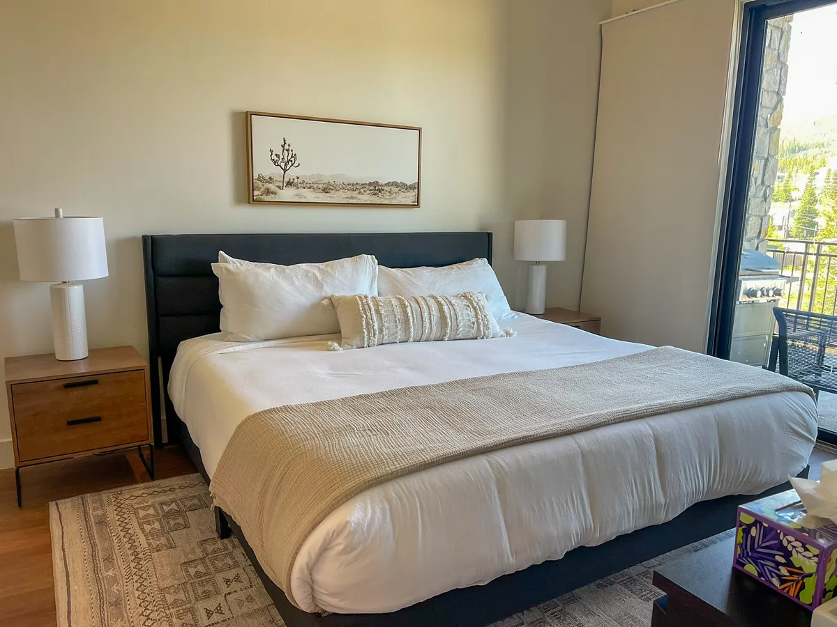 A stylish bedroom with a plush bed, soft gray bedding, bedside lamps, and art above the headboard. Ample natural light pours in through a window, creating a serene retreat perfect for restful nights.