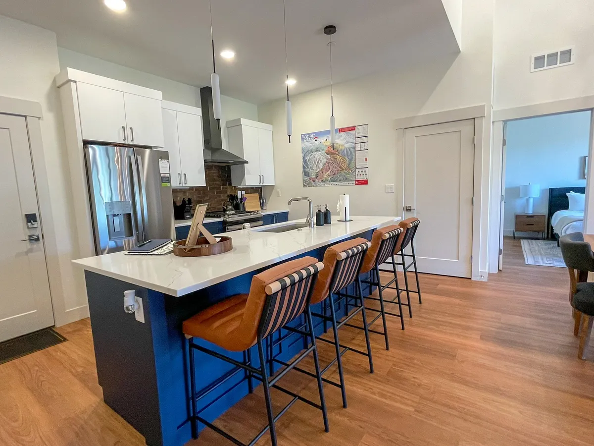 The image features a bright, stylish kitchen with a spacious island, modern bar stools, sleek stainless steel appliances, and tasteful mountain-inspired artwork. The space is well-lit and designed for both cooking and socializing, inviting guests to gather for meals or drinks.