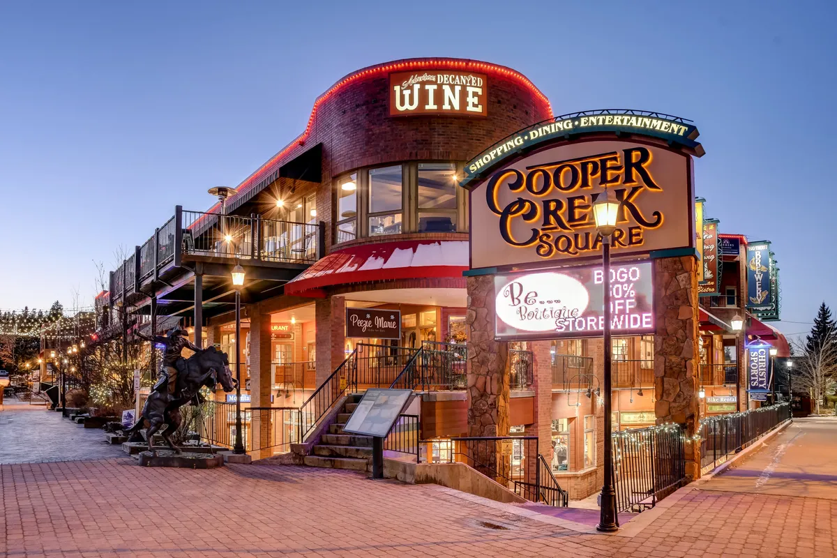 A lively exterior image of a shopping and dining complex, featuring the Cooper Creek Square sign, a wine shop entrance, outdoor seating, and festive lighting. The scene is bustling and welcoming, perfect for strolling, dining, and exploring local boutiques.