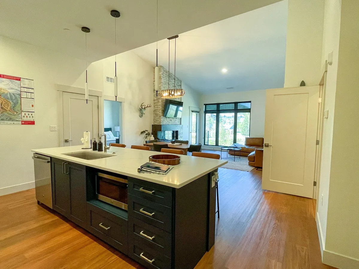 This image showcases a contemporary open-plan living area that includes a kitchen island, living space with large windows, and a striking fireplace under a stylish chandelier. The arrangement highlights the seamless connection between kitchen and living areas, with cozy yet modern furniture and plenty of natural light from the expansive windows.