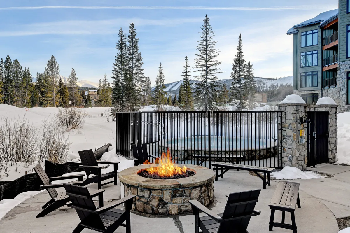 A communal outdoor area featuring a glowing fire pit surrounded by chairs near a bubbling hot tub, all set in a snowy landscape bordered by trees. The atmosphere is warm and sociable, inviting group gatherings or quiet moments by the fire.