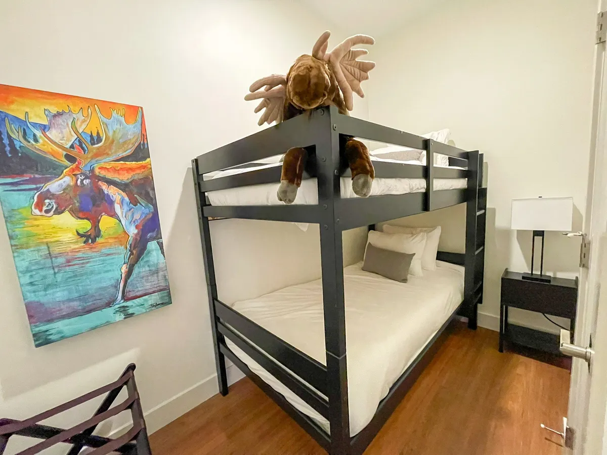 A playful bunk space with a twin-over-twin bunk bed, colorful moose artwork, a plush moose toy, and a lamp-lit nightstand. The scene is cheerful and inviting, ideal for kids or friends sharing an adventure.